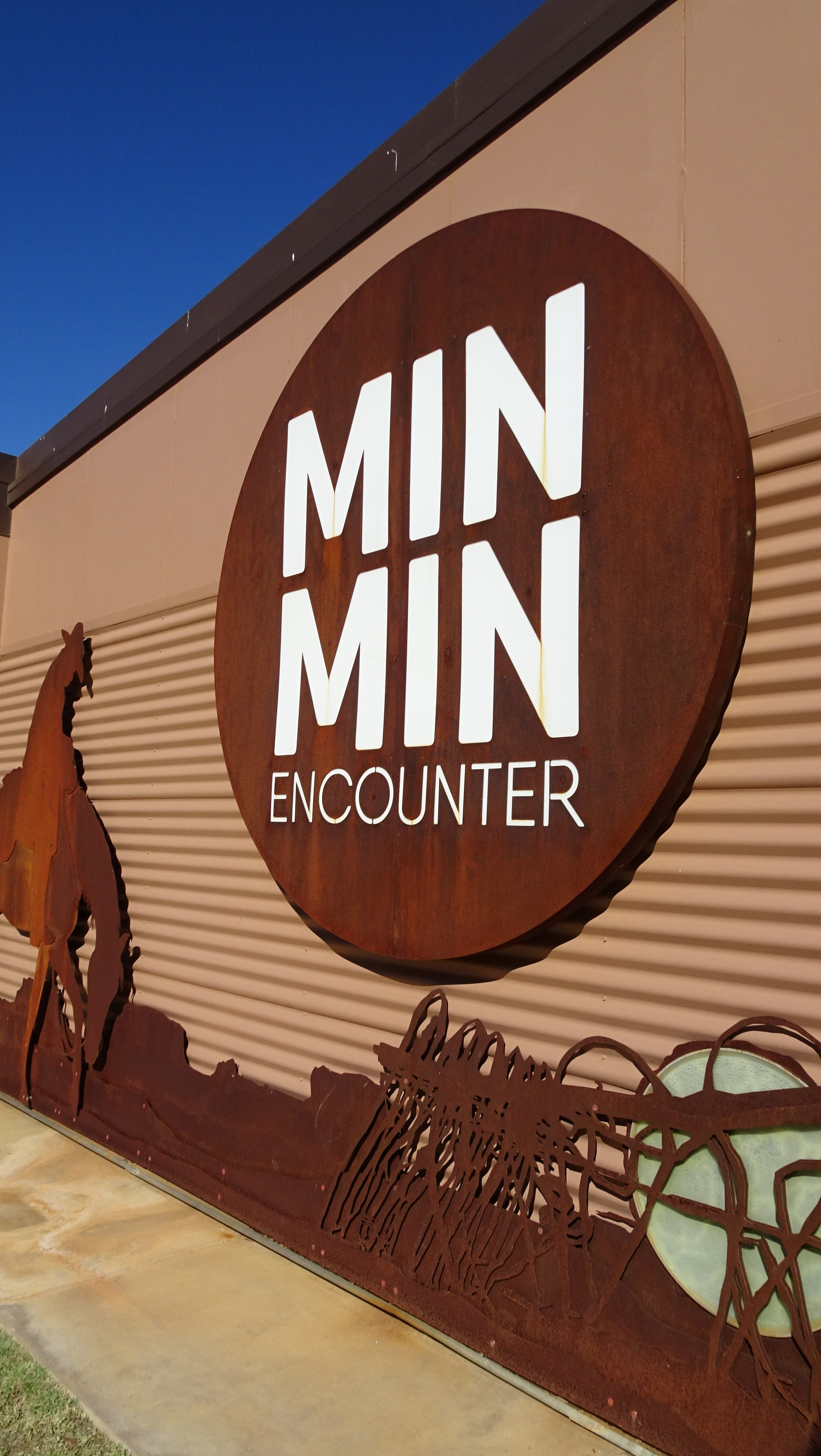 Exterior of Boulia's Min Min Encounter with signage, stockman on a grazing horse and lurking Min Min light